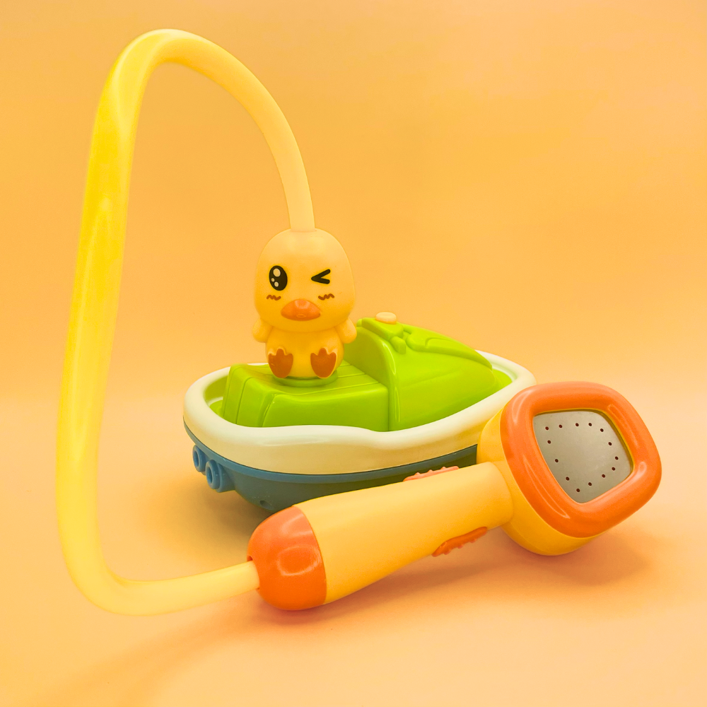 A children's toy water play set featuring a small yellow duck sitting on a green and blue boat, with a yellow hose and a yellow-and-orange handheld shower attachment, photographed on a warm peach background.