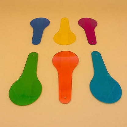 Six colorful plastic paddle-shaped tools in blue, yellow, magenta, green, orange, and teal, arranged on a warm beige background.
