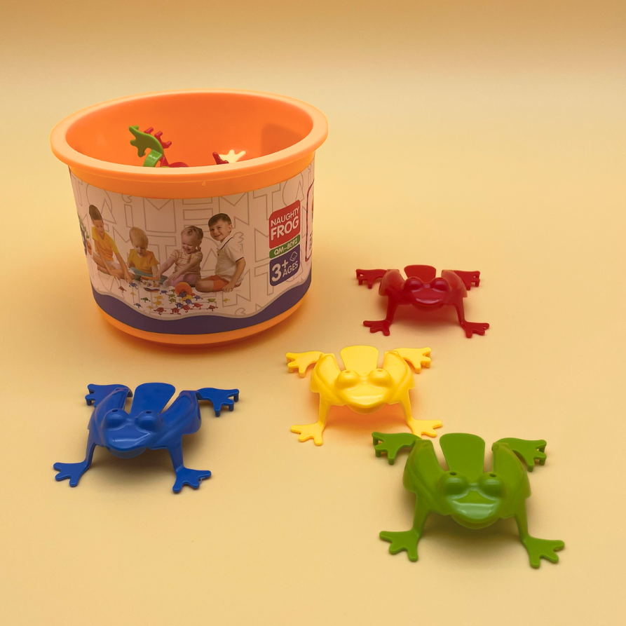 A children's toy set featuring four plastic frog figures in blue, yellow, green, and red, arranged around an orange bucket container with a printed label on a warm beige background.